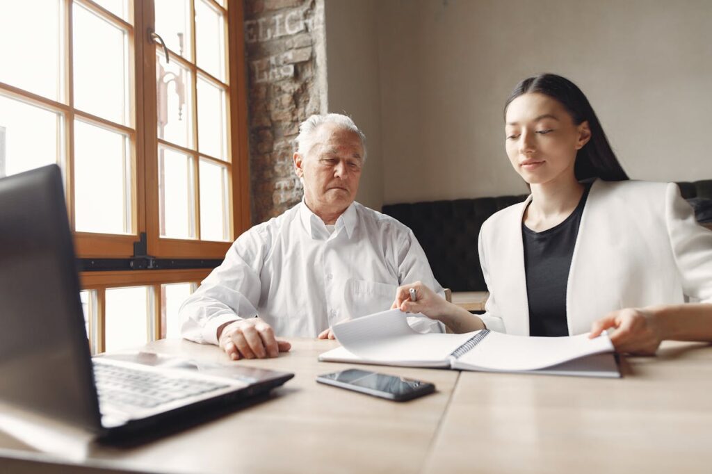 Focused elegant senior man and adult woman in smart casual outfit working on project with laptop and notes together in modern workspace